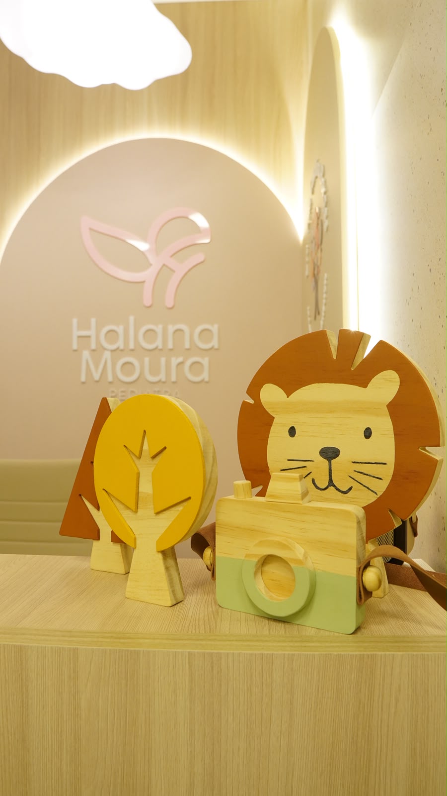 Recepção do consultório, com o letreiro Halana Moura Pediatra na parede e brinquedos de madeira sobre o balcão